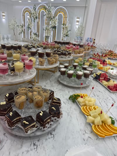 The image depicts a dessert and fruit buffet at a venue, likely for an event, in Mălăiești, Moldova. The table is laden with an array of sweets, including individual layered chocolate cakes, small glasses filled with layered desserts or mousses, and shot glasses containing colorful layered beverages or custards. There are also platters of fresh fruit, such as sliced melon and oranges, garnished with parsley and decorative picks. In the background, a decorative archway with floral arrangements and lighting suggests a festive or elegant setting. No people are visible in the frame. The overall environment appears to be indoors, with a marble-patterned tabletop and white walls.