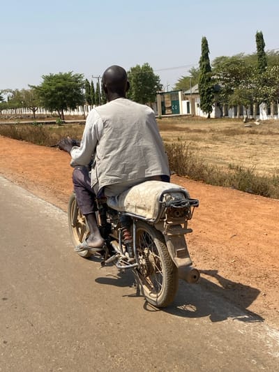 A dark-skinned male, bald, is depicted from the rear, operating a motorcycle on an asphalt road in Lapai, Nigeria. He wears a light grey long-sleeved garment and dark trousers, with a single light-colored sandal visible on his left foot. The motorcycle is an older, utilitarian model featuring a tan-colored seat cover, gold-colored spoked wheels, and a rear cargo rack; the vehicle appears dirty and worn. The asphalt road transitions to a reddish-brown dirt shoulder on the right, sparsely covered with dry, light-brown vegetation. In the midground, a line of green trees runs parallel to the road on the left, while a large, dry field extends to the right. The background features light-colored buildings with green roofs, a partially visible green entrance gate marked "CO," and tall, narrow dark green trees. Utility poles are also present. The scene is illuminated by bright daylight under a clear, light blue sky, casting distinct shadows on the road from the motorcycle and rider.