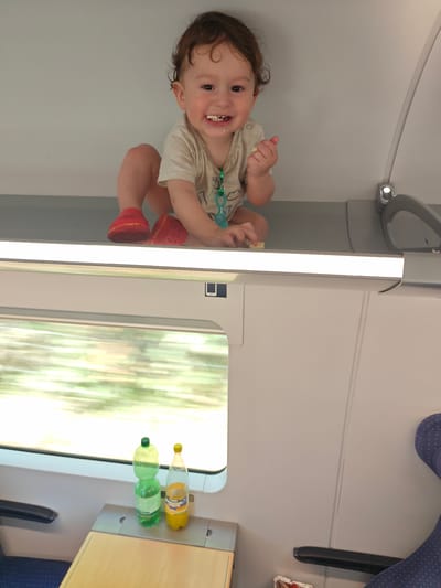 A toddler, with curly brown hair and a wide smile showing small teeth, is sitting on a shelf above a train window. The child wears a light-colored t-shirt and red shoes. In their right hand, the toddler holds a piece of food. The background is a plain wall, with a light fixture visible.

Below the shelf, a train window shows a blurred, green and yellow landscape, indicating the train is in motion. On a ledge beneath the window, two drink bottles are placed: one green bottle filled with a clear liquid, and a yellow bottle with an orange-colored liquid. A small wooden tray is also visible.

The scene appears to be indoors, likely within a train carriage. The lighting suggests it could be daytime. The toddler's expression is joyful. The location context is Mülheim an der Ruhr, Germany, which is a city known for its industrial history and location along the Ruhr River.