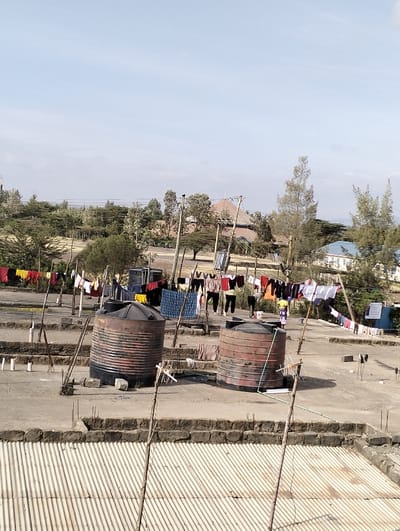 Two large, weathered, cylindrical water tanks sit on a flat concrete rooftop. Clotheslines strung between poles are heavy with colorful garments drying in the sun. In the background, trees and buildings dot the landscape under a partly cloudy sky. The foreground shows a corrugated metal roof, and the whole scene conveys a sense of rural life.