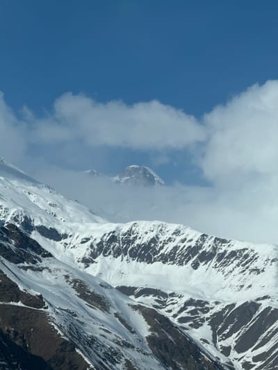 The scene depicts a snow-covered mountainous landscape, identified as being located in the city of Kanobi, Georgia. The upper third of the image consists of a clear, deep blue sky. Below this, a band of white cumulus clouds partially obscures the horizon. Through a gap in these clouds, a distant, prominent, rugged peak, heavily coated in snow, is visible, flanked by a lesser, partially cloud-enshrouded peak to its left. The mid-ground and foreground are dominated by multiple layers of snow-covered mountain ridges and slopes. The upper mid-ground mountains exhibit significant snow accumulation interspersed with exposed dark rock formations, particularly on steep inclines, creating a striped or patterned effect. The lower foreground slopes show a less continuous snow cover, revealing patches of dark, possibly rocky or barren earth between snowdrifts. No individuals, man-made structures, or signs of activity are present within the depicted environment.