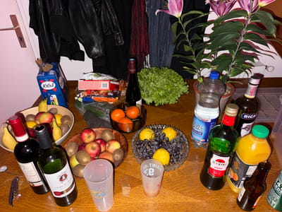 The image shows a wooden table laden with various items, suggesting a gathering or party. There are several bowls filled with fruit, including apples, kiwis, bananas, oranges, lemons, and grapes. Two bottles of wine, one with a red label and one with a green label (Lussac St. Emilion De Lussac), lie on their sides next to a corkscrew and some plastic cups. Other beverages include a bottle of Ricard, orange juice, a bottle of Ravini Rosso, and a bottle of beer marked "1664". There is also a bottle of water and a head of lettuce. A potted plant with purple flowers sits at the back of the table. In the background, a black leather jacket hangs next to a denim jacket. The location in Montreuil, France, is not discernible from the image.
