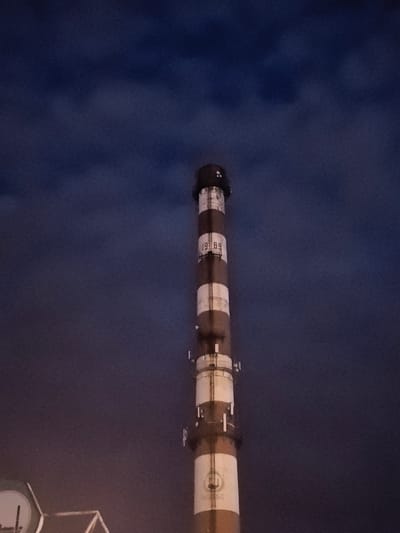 The image shows a tall, industrial smokestack against a dark, cloudy sky. The smokestack is cylindrical and painted with alternating white and red-brown bands. It has several sections with visible metal structures and what appear to be antennas or monitoring equipment attached to its sides. Near the top of the smokestack, there is a white rectangle with the numbers "19 89" clearly visible. Further down, there is a circular logo with some faint text beneath it. The bottom of the smokestack is partially obscured by some structures, one of which is a white circular object with a dark interior and a dark vertical line. The sky is a deep, moody blue with heavy cloud cover, suggesting it is either dusk, dawn, or a very overcast day. The overall atmosphere is industrial and somewhat somber. The presence of the smokestack and the industrial equipment indicates an industrial setting, likely a factory or power plant. The text "19 89" may represent a year, possibly related to the construction or operation of the facility. The location context provided is Ново-Талицы, Russia.