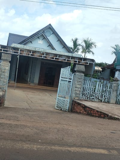 The image shows a building viewed from a street in Thôn 4, Vietnam. The building appears to be a church or religious structure, with a triangular roof, gray exterior, and a covered entryway supported by pillars. A gate made of metal bars stands partially open, leading to a paved driveway. The street in the foreground is unpaved and uneven. A partial view of a wooden fence and another structure is visible to the right of the building. Palm trees can be seen in the background. The sky is overcast with light clouds.
