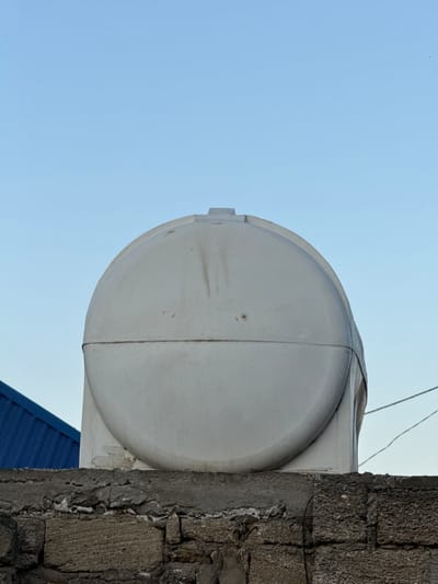 The image shows a low-angle view of a white, cylindrical water tank situated outdoors. The tank is positioned next to a rough, stone-like wall made of large, irregular blocks. A portion of a blue, corrugated roof is visible beneath the water tank and to the right of the wall. The sky above is clear and a pale blue, suggesting daytime. The overall scene appears to be residential or utilitarian, possibly on a rooftop or elevated platform. There is no visible activity or people in the frame. The sunlight casts soft shadows, indicating it is likely daytime, but the angle doesn't suggest a specific time like sunrise or sunset. The location cues, such as the style of the wall and roof, along with the clear sky, are consistent with an outdoor setting in Mastaga, Azerbaijan. No text is visible in the image.