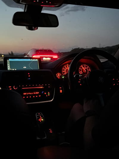 The image depicts an interior perspective from a BMW vehicle at dusk or night, looking forward through the windshield. The vehicle's dashboard, instrument cluster, and center console controls are illuminated in red. A driver's hand, wearing a watch, rests on the black multi-function steering wheel, which prominently displays a BMW emblem. A passenger's leg and arm are partially visible in the bottom right.
The instrument cluster features two main analog gauges, illuminated in red, alongside a smaller gauge to the left. A central infotainment screen displays a navigation map with a highlighted route, showing a battery charge of "93%" and the time "20:49". Below the screen, the climate control and audio system display temperature settings of "19.5°" and "20.0°". The illuminated electronic gear selector indicates "P" and "M/S" options with a green light suggesting an engaged drive mode. The rearview mirror is visible at the top center, reflecting the ambient light.
Outside the vehicle, a red car is visible ahead on what appears to be a highway or multi-lane road. Its distinct, horizontal brake lights are illuminated. The sky above the horizon shows residual twilight, with dark, indistinct landforms or structures in the distance. The license plate of the car ahead is unreadable.
Based on the visual evidence, the car is likely stationary or moving slowly. The metric temperature units and vehicle type are consistent with European use. However, there are no specific visual cues or identifiers within the image (such as recognizable landmarks, legible road signs, or discernible license plate details) to forensically confirm the precise location as Mursalevo, Bulgaria.