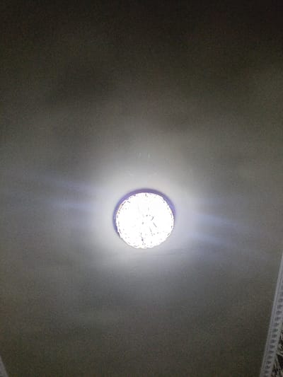 The image displays an interior scene in Bulungur, Uzbekistan, focused on a circular light fixture mounted centrally on a light grey ceiling. The fixture emits intense white light, causing central overexposure and visible light rays extending outwards. The outer perimeter of the light fixture reveals a textured or decorative pattern. The surrounding ceiling surface is plain light grey with minor dust particles visible. A patterned textile or decorative border, featuring dark designs on a lighter background, is partially visible along the lower right edge of the frame. No individuals or dynamic actions are present.