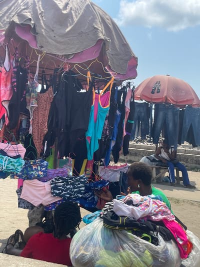 An outdoor market scene in Aiyetoto-Asogun, Nigeria, captured during daylight under a bright blue sky with scattered white clouds.

In the foreground and central-left, a makeshift clothing stall is visible, characterized by a large, worn, beige-brown tarpaulin or fabric cover atop a metal frame, with a layer of pink fabric visible underneath the primary cover. Various items of clothing, predominantly women's and children's swimwear and underwear, hang from numerous wire hangers. These items display a range of colors including black, turquoise, pink, and brown, along with patterns such as sharks and animal prints. Folded items are also arranged on lower display sections of the stall.

To the immediate right of the stall, an adult female with short, dark hair, wearing a green t-shirt and silver earrings, is observed examining clothing. Her right hand holds a folded white garment with pink floral patterns, while her left hand is positioned near other merchandise. Next to her, a large transparent plastic bag filled with assorted clothes rests on the ground. Partially visible in the lower foreground, another individual with dark, braided hair, wearing a red shirt, is seated low to the ground behind the clothing stall, facing away from the viewer.

In the midground-right, a faded red Coca-Cola branded umbrella provides shade. Beneath this umbrella, several pairs of blue denim jeans are suspended from a display structure. Seated on a low wooden bench beneath the Coca-Cola umbrella is an adult individual, partially obscured by the hanging jeans, wearing a white t-shirt and blue track pants.

The ground surface appears to be dirt or sand. A low concrete wall or barrier is visible in the background, behind the denim jeans display.