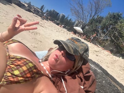 The image is of a woman lying on the beach in Coolangatta, Australia, wearing a bikini and a baseball cap. She is lying on her back with her head near a paved area that separates the beach from the street. She is wearing sunglasses and has earbuds in her ears. She is making a kissy face at the camera and holding up her hand in a peace sign. In the background, there are trees, buildings, and other people on the beach. The sky is blue.