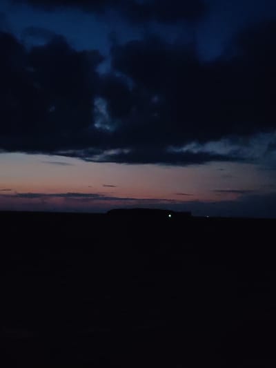 The image depicts a dark, twilight landscape with heavy, dark clouds dominating the upper portion of the frame. Below the clouds, a faint band of pink and purple hues suggests a fading sunset. The horizon is marked by a dark, silhouetted landmass, possibly a hill or a distant cityscape. In the foreground, the land is entirely black, suggesting deep shadow or night. A single, faint green light is visible on the silhouetted landmass. The scene is devoid of any visible people, animals, or specific structures that would allow for identification of the location within Larionovo, Russia. The overall impression is one of a moody, atmospheric natural setting at dusk or just after sunset.