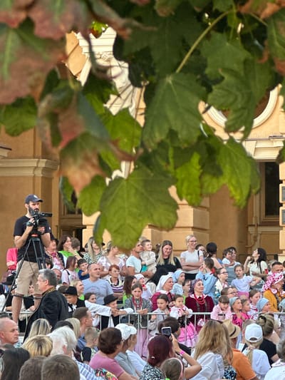 A medium shot captures a crowd of people gathered outdoors, with a man operating a video camera on a tripod in the foreground. The man, wearing a dark t-shirt and a baseball cap, is positioned to the left of the frame. Behind him, a diverse group of individuals, including children and adults, are visible. Many are looking towards the right, suggesting they are observing an event. Some are dressed in traditional folk costumes, characterized by colorful embroidery and headscarves. The background reveals an ornate building with classical architectural features, including columns and arched windows. Lush green leaves from a nearby tree partially obscure the view of the building and the sky, creating a dappled lighting effect. The scene appears to be a cultural event taking place in Nowy Sącz, Poland.