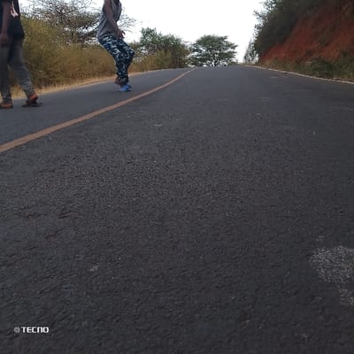 The image displays a low-angle perspective of an asphalt road extending into the distance, curving gently to the left and appearing to go uphill. The road surface is dark gray with a visible textured quality. A single, faded yellow line runs along the left edge of the road.

Two individuals are partially visible on the left side of the road. The person in the immediate foreground, on the far left, has their back mostly to the camera, wearing dark pants and light-colored sandals, appearing to be walking away. Further up the road and slightly to their right is a second person, wearing a gray sleeveless top and camouflage-patterned pants (blue, white, and dark gray), with light blue sneakers. This person is captured in a mid-stride or dynamic pose, with one foot slightly lifted.

The surroundings consist of natural vegetation. To the left of the road, there are dry, sparse trees with light-colored trunks and branches, and brownish ground cover. To the right, there is a reddish-brown dirt embankment or hill, partially covered with green foliage at its crest. The sky above is light and somewhat hazy, suggesting either early morning or late afternoon. The overall lighting is soft, with no harsh shadows.

In the bottom left corner of the image, a white watermark is visible, which reads "© TECNO".