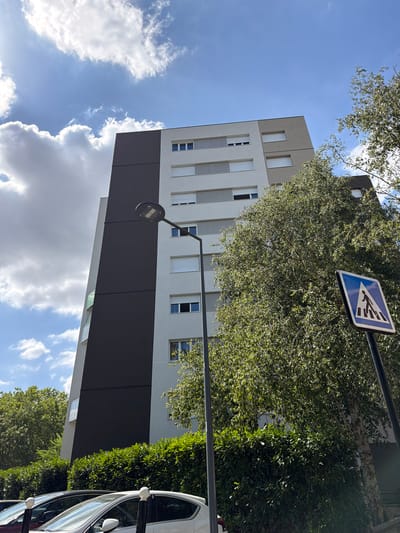 A low-angle shot captures a modern, multi-story apartment building in Évry-Courcouronnes, France, against a backdrop of a bright blue sky dotted with white clouds. The building features a distinctive facade with a large dark gray section contrasting with lighter gray and white panels. Rows of windows with closed white blinds are visible on the lighter sections. A tall, dark lamppost stands in the foreground, with a pedestrian crossing sign mounted on a pole to its right. Lush green foliage from trees and hedges partially obscures the lower part of the building and the street. Several cars are parked in front of the hedges, with their roofs and windshields visible. The scene appears to be an urban residential area on a sunny day.