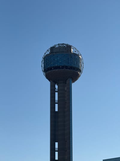 A low-angle shot captures the Reunion Tower in Dallas against a clear, blue sky. The tower's distinctive geodesic sphere at its top is partially illuminated. A concrete structure extends horizontally from the sphere. The sky is cloudless and uniform in color. A partial view of another building peeks in at the top right corner.