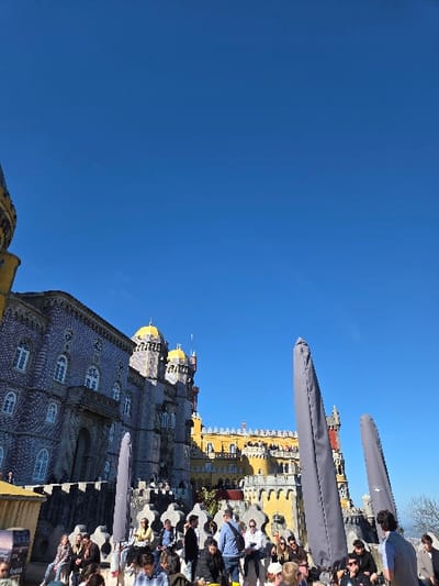 The image displays a daytime scene at the Pena Palace in Sintra, Portugal, specifically within the civil parish of Santa Maria e São Miguel, São Martinho e São Pedro de Penaferrim. The upper two-thirds of the frame are dominated by a clear, unclouded blue sky.
The Pena Palace occupies the midground and background, characterized by distinct architectural sections. A large section on the left features blue and white patterned azulejo tiles with multiple white-framed windows. Adjacent to this, the palace transitions into sections with yellow walls, adorned with yellow-gold domes and turrets. A red-colored tower is visible further in the background on the right. Numerous individuals are visible on the upper balconies and battlements of the palace structure.
In the foreground and lower midground, a crowd of approximately 30 to 40 people is gathered on an elevated platform or terrace. The individuals are engaged in various actions, including standing, conversing, and observing the surroundings. Their attire is casual. Two prominent objects in the midground are large, gray, folded outdoor umbrellas, positioned vertically and partially obscuring parts of the palace. A low, ornate stone wall or barrier runs across the foreground, indicating the edge of the viewing platform. A dark, rectangular display stand or sign is visible in the lower-left quadrant of the image. The entire scene is illuminated by strong natural daylight.