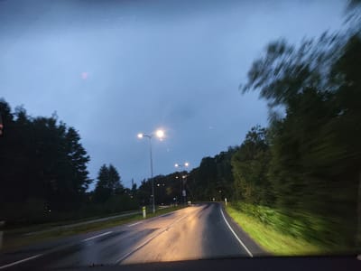 The image depicts a road at dusk or dawn in Harkujärve, Estonia. The road is wet, suggesting recent rain, and is illuminated by streetlights and the headlights of a vehicle from which the photo appears to be taken. The road curves to the right and is bordered by trees on both sides. The sky is a pale blue with some clouds. The overall impression is of a quiet, somewhat gloomy evening drive.