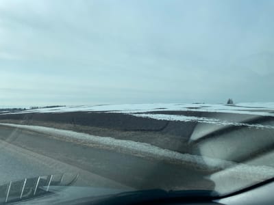 The image depicts a rural scene, likely a winter landscape. The foreground shows a road, partially covered in ice or snow, viewed from inside a vehicle, indicated by the dashboard and reflections on the windshield. Beyond the road is a field with patches of dark soil visible, contrasting with areas covered in snow. In the distance, the horizon line is visible, with more fields and scattered trees. The sky is overcast and gray, typical of winter weather. The location is given as Bol'shaya Sosnova, Russia, but without additional cues, no specific recognizable features from the location are present in the image. There are no people visible in the frame.