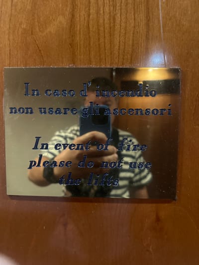 A metallic plaque is affixed to a wooden surface, displaying text in Italian and English. The Italian text reads, "In caso d'incendio non usare gli ascensori," which translates to "In case of fire do not use the elevators." The English text below it says, "In event of fire please do not use the lifts." A reflection on the plaque shows a person holding a blue smartphone and wearing a striped shirt. The background appears to be a wooden door or wall. The scene is likely in San Martino Buon Albergo, Italy, given the Italian text on the sign.
