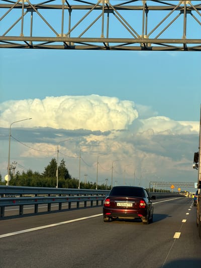The image depicts a highway scene with a large, white cumulonimbus cloud formation dominating the sky. The viewpoint is from a vehicle traveling on the highway, looking forward. A dark red sedan is in the foreground, in the right lane, its license plate reading "E 125OK 716". The highway has a metal guardrail on the left and a concrete barrier on the right, with streetlights lining the road. Trees and utility poles are visible beyond the guardrail. In the distance, traffic signs and a yellow sign with an exclamation mark are present. The sun appears to be low, casting long shadows and illuminating the clouds with a warm light. The location is identified as the city of Egor'evo, Russia.