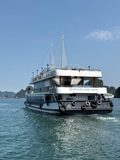 A large, modern luxury cruise ship is depicted sailing on calm, blue-green waters under a clear, bright sky. The vessel, predominantly white with black accents along its hull, is seen from a side-rear angle, moving from left to right and leaving a distinct wake.

The ship features multiple decks with large windows and exterior stairs leading to its upper levels. Decorative masts or antennae with numerous rigging lines rise from the top deck, adorned with a string of small, colorful pennants. Along the black portion of the hull, several dark, circular mooring fenders (likely repurposed tires) are attached. Clearly visible in green lettering on the ship's side is the text "HERITAGE LUXURY CRUISE," accompanied by a stylized logo.

In the background, under a vast expanse of light blue sky with a single, wispy white cloud, are several lush green limestone karsts or islands. These geological formations are typical of the scenic landscapes found in regions such as Halong Bay or Lan Ha Bay in Vietnam, consistent with the Tuan Chau Ward context. The water's surface subtly reflects the sunlight, showing gentle ripples.

The scene captures a serene daytime setting, likely late morning or early afternoon, characterized by clear and sunny weather, evoking a sense of leisure and natural beauty. No individuals are distinctly visible on the ship's decks or through its windows. The overall image portrays a peaceful cruise experience amidst a picturesque natural environment.