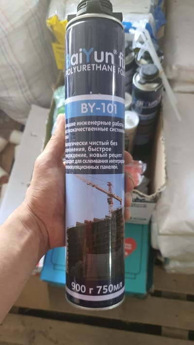 A hand holds a can of polyurethane foam labeled "BaiYun" and "BY-101" against a background of construction materials. The can is predominantly dark blue with white and light blue text and features an image of a construction crane and building. The text on the can is in Russian and describes the product as high-quality engineering work, environmentally clean, fast curing, and suitable for bonding integrated insulation panels. The bottom of the can indicates "900g 750ml". The scene appears to be in a warehouse or storage area.