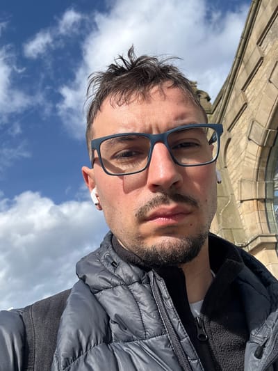 A man is pictured in a selfie, wearing glasses and a dark gray puffer jacket with a black fleece collar. He has short brown hair and a dark beard. He has what appear to be white wireless earbuds in his ears. The background includes a blue sky with white clouds, and the ornate stonework of a building. The location is most likely in Sheffield, United Kingdom, given the architecture and the prompt.