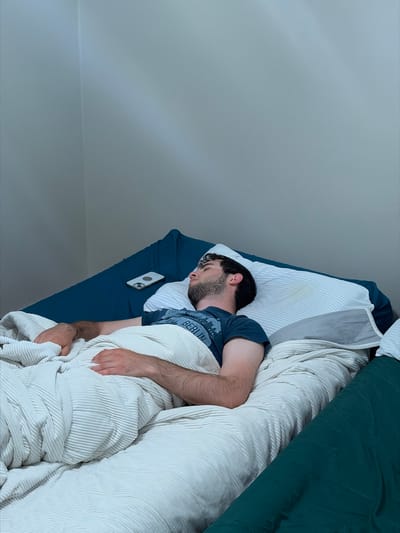The image captures a man sleeping or resting in a bed, viewed from a high angle looking down. He is lying on his right side, facing towards the left of the frame, with his head on a white pillow. He has short, dark hair and a beard. He is wearing a dark blue t-shirt with the word "BERLIN" and a cityscape graphic visible on the front.
The bed features a dark blue fitted sheet. A thick, ribbed white blanket or duvet covers most of his body, while another dark green duvet or blanket is partially visible at the top right, folded on the bed. A white smartphone with a dark camera module is placed on the blue sheet next to his head.
The lighting in the room appears dim, suggesting it might be early morning, late evening, or simply a darkened room. The background consists of plain, light gray walls, with a subtle line indicating a wall or ceiling join at the bottom left. The overall scene depicts a quiet, indoor bedroom setting. The man appears to be in a state of peaceful slumber. The location context provided for this media file is Franklin Township, United States of America.