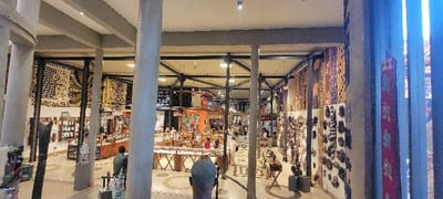 This image depicts the interior of a large, multi-level retail establishment, identified as a craft or souvenir market, located in Mto wa Mbu, Tanzania. The expansive space features a high ceiling supported by numerous tall, cylindrical concrete pillars and exposed grey and black structural beams.

The walls are extensively decorated with a variety of patterned textiles, large tapestries, and mounted artisanal items such as wooden masks and plaques. Merchandise is displayed across various setups: glass-topped wooden counters and tables occupy the central area, showcasing smaller items like jewelry, carvings, and cultural artifacts. On the left, shelves hold packaged goods, and large wooden sculptures, including a tall, dark abstract piece and several colorful carved figures, are arranged on pedestals. The floor consists of light-colored tiles with darker circular patterns.

Approximately 10-15 individuals are present within the market, observed in various states of browsing or walking through the displays. One individual's head is prominently featured in the foreground, facing away from the camera, wearing a grey shirt. The market is brightly illuminated by overhead artificial lighting, supplemented by natural light filtering through vertical, somewhat obscured window panels on the right side of the frame, which show glimpses of colorful patterns.