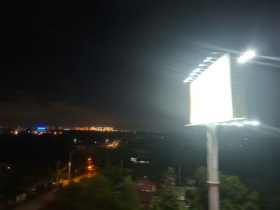 This is a night-time photograph taken from a high vantage point, likely a building or overpass, overlooking a city at dusk or night. The foreground is dominated by a brightly lit billboard on the right side of the frame, its surface glowing white and illuminated by rows of lights along its top edge. A tall, slender pole supports the billboard. In the middle ground, a cityscape unfolds with various buildings and structures. On the left, a building with blue, illuminated lettering is visible. Further into the distance, a line of buildings is lit with warm, yellow and red lights, suggesting a commercial or residential area. The sky above is dark, with some faint clouds visible. The overall scene depicts an urban environment at night, with artificial lights creating a contrast against the dark sky. The lighting suggests that the photograph was taken after sunset. There is no discernible weather activity. The visual information provides cues for a city location, and the provided location context of Ho Chi Minh City, Vietnam, aligns with the dense urban lighting and architecture. No specific activities or events are apparent, as the image focuses on the general landscape and static elements.