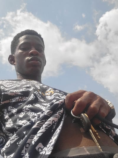 An outdoor, low-angle, head-and-torso shot depicts a dark-skinned male adult positioned against a clear blue sky with scattered white cumulus clouds, located in the city of Nwofia, Nigeria. The individual looks directly towards the camera with a neutral expression. He wears a dark short-sleeved shirt with a repeating pattern of white text. His right hand, visible in the lower right portion of the frame, holds a set of keys. The keys include at least two metallic keys and a black plastic key fob, all attached to a silver-colored key ring and a dark lanyard. A metallic watch band is present on his right wrist.