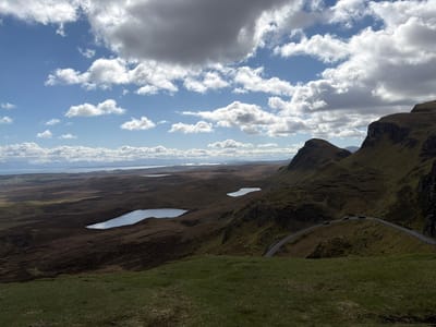 The image depicts a broad, undulating landscape in Portree, United Kingdom, under a sky filled with scattered cumulus clouds. Three lakes are visible in the middle ground, reflecting the sky. A winding road traverses the right side of the frame, with four cars traveling on it. The terrain is a mix of dark brown and green vegetation, with some areas appearing rocky. The foreground is predominantly green grass. In the distance, a body of water with land on the opposite shore is visible.