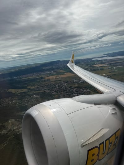 An aerial view from an airplane window shows a cloudy sky above a landscape of green forests, fields, and a city. The airplane's engine and wing are visible in the foreground, with the "Buzz" logo emblazoned on the side of the engine and the wingtip. The city appears to be located near a body of water, possibly the Black Sea. The terrain is varied, with hills and flatter areas. The time of day is daytime, given the illumination. The scene is static and shows no people or animals. The specific location in Pripek, Bulgaria, cannot be identified from the image alone.