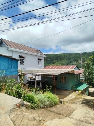 The image depicts a rural residential area in Xuân Sơn, Vietnam, on a cloudy day. In the foreground, a cracked concrete pathway leads to a yard with lush greenery. Two houses are visible: a larger, white, two-story house with a brown tiled roof on the left, and a smaller, teal-colored house with a red corrugated metal roof on the right. A blue structure is partially visible to the left of the white house. A white minivan is parked under a carport attached to the teal house. In the background, rolling green hills covered in trees rise under a sky with scattered clouds, and a cell tower stands prominently on one of the hills. Overhead, multiple power lines crisscross the frame.