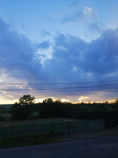 The image captures a twilight sky with dramatic clouds, tinged with hues of blue and grey, over a landscape in Fălticeni, Romania. The horizon is lined with a dark silhouette of trees. Three horizontal power lines stretch across the mid-section of the image. Below the trees, a dark fence stretches across the scene, with some green tarp visible behind it. A paved road occupies the foreground, fading into shadow. The scene appears quiet and devoid of human activity, evoking a sense of calm as day transitions into night.