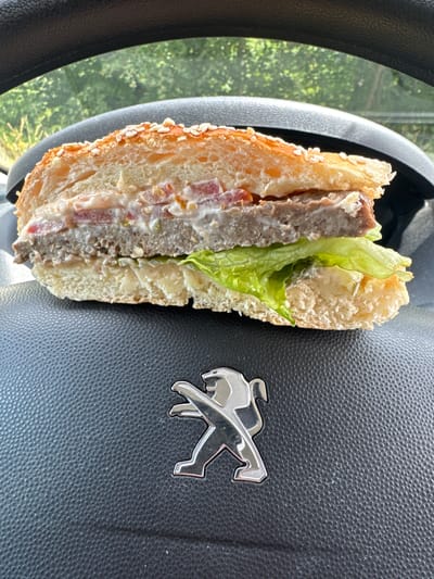 A half-eaten burger rests on the steering wheel of a car. The burger is cut in half, revealing the layers of lettuce, tomato, and a thick meat patty. The top bun is sprinkled with sesame seeds. The steering wheel is black and textured, with a silver Peugeot logo in the lower right corner. The background is a blur of green trees and foliage, suggesting the car is parked outdoors. The image was taken in Jülich, Germany, on a sunny day.