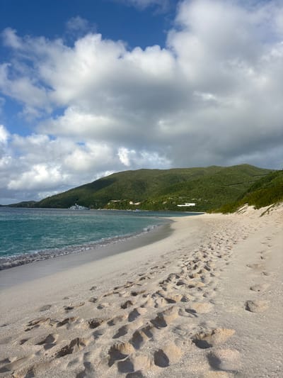 The image depicts a tropical beach scene in the vicinity of Olde Yard Village, British Virgin Islands. In the foreground and midground, a wide, light-colored sandy beach extends from the lower right, curving gently to the left where it meets the ocean. Numerous human footprints are deeply impressed into the sand, forming visible trails along the beach. To the left, the ocean displays clear, turquoise water with small, white-crested waves breaking softly on the shoreline.
The background is dominated by lush, green, densely vegetated hills that rise directly from the coastline. Several white structures, appearing to be buildings, are interspersed among the foliage on the hillsides. Anchored or moored in the distant water, near the base of the hills, are multiple vessels, including a large white superyacht and at least two smaller white boats. The sky above is partly cloudy, featuring patches of clear blue interspersed with large white and grey cumulus clouds. No individuals are visible on the beach or in the water.