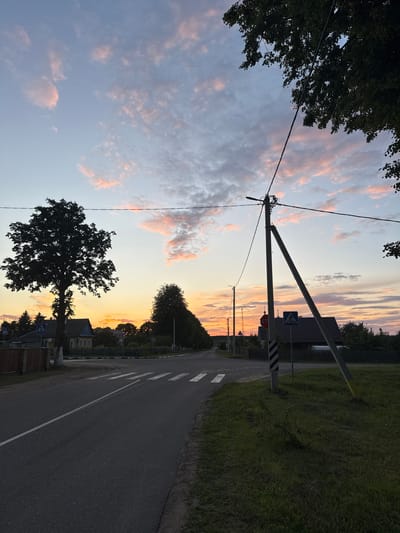 The image depicts a rural road in the Neman Sel'skiy Sovet, Belarus, at sunset. The sky is a gradient of pale blue at the top, transitioning to hues of pink and orange near the horizon, with scattered clouds catching the fading light. A paved road with a white line marking the center runs from the foreground towards the background, curving slightly to the right. A pedestrian crossing is marked on the road. On the left side of the road, a large, silhouetted tree stands prominently, with a fence and a yellow house visible behind it. Further down the road on the left, there's a pedestrian crossing sign. On the right side of the road, a utility pole with wires stretches across the frame, leaning slightly. Another utility pole is visible further down the road, and to its right, houses and trees form the background. The overall scene is one of quiet twilight in a rural Belarusian setting. There are no people visible in the image.