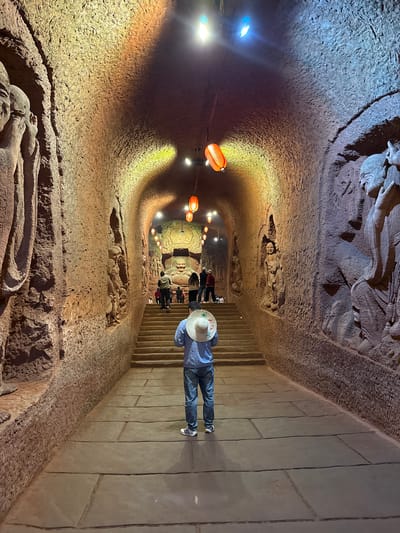 This image depicts an underground or cave passage, likely a grotto, adorned with intricate relief carvings, located in Shizhong, China. The scene is illuminated by artificial lighting, creating a dramatic interplay of warm and cool tones on the carved surfaces.

In the immediate foreground, a person stands on a paved walkway, seen from behind, wearing a light blue long-sleeved shirt, blue jeans, white sneakers, and a wide-brimmed straw hat. They are positioned near the top of a descending staircase, looking down into the curving tunnel.

The passage itself is tunnel-like, with both sides and the ceiling covered in detailed relief carvings. These carvings depict various figures, likely Buddhist deities, historical characters, or scenes, sculpted directly into the textured, earthy rock. The walls exhibit a rough, chisel-marked appearance, indicating their hand-carved nature.

Several orange, accordion-style lanterns hang from the ceiling at intervals, emitting a soft, warm light. Additional brighter, cooler-toned lights are embedded in the ceiling, illuminating different sections of the carvings and creating highlights and shadows that emphasize their depth and detail. Further down the tunnel, several other people can be seen, suggesting the site is a popular attraction.

The activity taking place is exploration or sightseeing within this historical or cultural grotto. The setting is clearly indoors and underground, with no visible cues for time of day or weather. No visible text is present in the image.