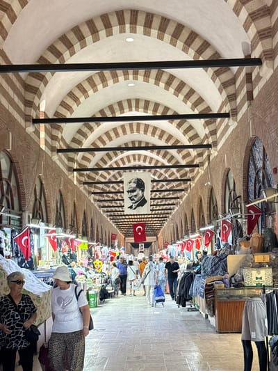 The image depicts the interior of a market hall in Edirne Merkez, Turkey, characterized by its arched ceiling with alternating brown and white stripes and dark support beams. The hall is lined with stalls selling various goods, predominantly clothing and textiles, adorned with numerous Turkish flags. Several individuals are present, walking through the market, browsing stalls, and engaged in shopping. In the center of the hall, a large black and white portrait of Mustafa Kemal Atatürk hangs prominently. The flooring appears to be paved stone. The overall impression is that of a bustling, traditional marketplace.