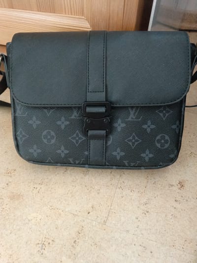 This is a close-up, eye-level shot of a dark grey messenger bag with a Louis Vuitton-inspired monogram pattern. The bag is the primary subject, filling most of the frame. It appears to be made of textured leather and features a black buckle closure and strap. The background consists of a light-colored, speckled surface, likely a floor, and a wooden surface with vertical slats positioned above the bag. The lighting suggests an indoor setting, possibly a home or a retail environment, with no strong indications of the time of day or weather. No people are visible, and no text is present on the bag or in the background. The scene is static, focusing solely on the presentation of the bag. The bag itself displays the iconic Louis Vuitton monogram in a slightly darker shade of grey, creating a subtle yet recognizable pattern. The overall aesthetic is modern and luxurious, with a focus on the design and material of the bag.