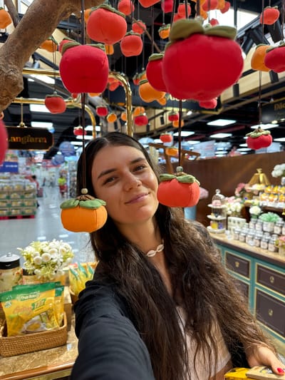 In a retail setting that appears to be a store in Phu Thuy, Vietnam, a young woman with long, dark, wavy hair is the central subject. She is positioned in the foreground, looking towards the camera with a slight smile. She is wearing a white top and a dark jacket. A distinctive shell necklace adorns her neck.
The background depicts a brightly lit store interior with shelves stocked with various products. Hanging from the ceiling are numerous decorative items resembling persimmons, some red and some orange, made of fabric. These are suspended by strings, creating a festive atmosphere. Behind the woman, to her left, a basket contains several yellow packages with Vietnamese text, likely food or drink items. To her right, shelves are filled with jars and bottles. A sign in Vietnamese, "Langfarm," is visible in the mid-ground. The overall impression is that of a market or a specialty store, possibly selling local produce or artisanal goods. The lighting is artificial and bright, typical of an indoor retail space.