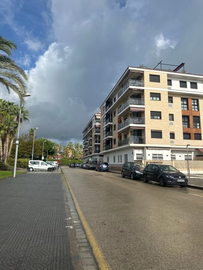 The image displays a street scene in Pineda de Mar, Spain, captured under a sky exhibiting a mix of bright blue patches with white clouds and expansive dark grey, heavy cloud formations.

In the foreground, a dark grey paved sidewalk with a distinctive circular and linear pattern occupies the left side, transitioning into a grassy area dotted with mature palm trees. A silver street lamp with a white fixture stands prominently on the left. Adjacent to the sidewalk on the right is an asphalt road marked by a yellow curb line.

Moving into the midground, several vehicles are parked along both sides of the road. On the left, a white panel van is parked perpendicular to the street, followed by a lighter-colored hatchback in a parallel parking spot. Further down the road, more vehicles are partially obscured by additional palm trees and street lamps. On the right, two multi-story residential buildings with light beige facades dominate the scene. These buildings feature multiple balconies with glass railings and retractable awnings, typically striped in red and white. The closest building also incorporates sections of darker brown brickwork or tiling into its exterior. A low, white ornamental fence runs along the base of the buildings. Directly in front of these structures, a line of cars is parked parallel to the curb, including a dark grey hatchback, a blue hatchback, a dark grey sedan, and a black hatchback or small SUV displaying a German license plate "DDG MG 503."

The background reveals more palm trees and distant green foliage or hills under a slightly brighter patch of sky, contrasting sharply with the darker clouds directly overhead. No individuals are present in the scene, and no active events or interactions are observed.