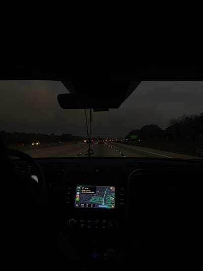 A forensic description of the image reveals a nighttime or heavily overcast daylight scene from the interior of a vehicle traveling on a multi-lane highway, consistent with the Boca Raton, Florida, United States area. The car's navigation system displays a map and indicates a destination "17 mi" to "Exit 07-08-09 / Southern Blvd," a major road in Palm Beach County, just north of Boca Raton.
The driver's right arm and hand are partially visible on the steering wheel on the left side of the frame. The vehicle's dashboard and center console feature an illuminated infotainment screen with an active navigation map. Below the screen, several controls are visible, including backlit buttons and rotary knobs emitting a blue light. Centrally positioned overhead, a rearview mirror is present, from which a dark, cross-shaped object hangs by a string.
Through the windshield, the multi-lane highway extends forward. Distant vehicle lights, both red tail lights and white headlights, indicate traffic flow. Reflective road markers delineate the lanes. Along the right side of the highway, a green overhead road sign is partially visible amidst silhouetted trees. The sky is uniformly dark gray and overcast.