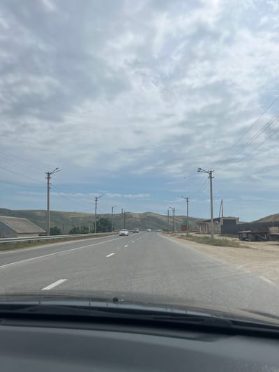 The image shows a highway on a cloudy day, with rolling hills in the background. The road is asphalt and appears to be in good condition, with white lane markings. There are several cars on the road, and utility poles with power lines line the sides of the highway. A building with a gray roof is visible on the left side of the road, and a red truck is parked on the right side. The sky is filled with gray and white clouds, indicating overcast weather. The overall scene suggests a rural or suburban setting outside of Karabudakhkent, Russia.