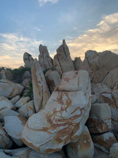 The image depicts a rugged landscape dominated by large, weathered rock formations and sparse vegetation. The rocks are varied in shape and size, with some appearing sharp and jagged, while others are more rounded. Their surfaces display a texture of rough stone with natural striations and patches of reddish-brown coloration, suggesting mineral deposits or weathering patterns. Green foliage, appearing as small bushes and dry grasses, is visible in crevices and at the base of some rock clusters, indicating a somewhat arid environment. The sky above is a blend of pale blue and muted orange and yellow hues, with wispy clouds, suggesting either dawn or dusk. The overall impression is one of a natural, undisturbed geological area, possibly a coastline or a rocky terrain. The location is specified as Thuận Thành, Vietnam.