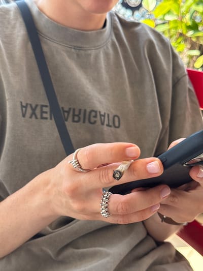 The image shows a close-up of a person's hands and torso, wearing a grey t-shirt with text that appears to read "ADIAS VEXA". One hand holds a lit cigarette, with ash visible at the tip. The person is wearing two rings: one on the index finger with a barbed wire design, and another on the middle finger with a series of small spikes. The person is also holding a dark-colored smartphone. The background is blurred, but suggests an outdoor setting with greenery and possibly a red chair. The lighting indicates it is daytime. The overall impression is casual and relaxed.