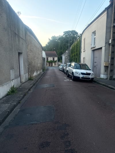 This image depicts a street in Longpont-sur-Orge, France, characterized by its residential architecture and parked vehicles. The street itself is paved with a dark asphalt surface with reddish aggregate. A prominent feature is a white Skoda Yeti, license plate CY-049-SP, parked on the right side of the road. Several other cars are visible parked behind it, forming a line.

The buildings lining the street exhibit a mix of architectural styles. On the left, there is a textured, grey stucco building with a stone facade and a narrow doorway. Further down the street, a more traditional building with a red-tiled roof and stone walls is visible. On the right, a light grey building with a large wooden garage door and a closed shuttered window dominates the foreground. Another building with ivy-covered walls and windows is visible further down the street on the right.

The environment appears to be a quiet, residential street, possibly taken in the late afternoon or early evening, given the soft light and clear sky. There are no people visible in the image. The scene is static and orderly, with the parked cars and houses suggesting a typical day in a French town. Utility poles and overhead wires are also present, indicating the presence of essential services. The overall impression is one of tranquility and everyday life.