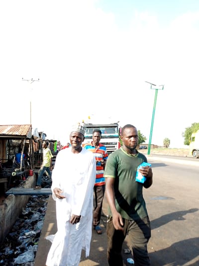 In Gadao, Nigeria, three men are captured in a photograph walking on a paved road. The man in the foreground, dressed in a dark green t-shirt and dark pants, is holding a small, light blue object, possibly a packet of tissues or wipes. He has a neutral expression and appears to be looking towards the camera. To his left, another man is walking, wearing a white traditional robe and cap, his gaze also directed forward. Behind them, a third man in a horizontally striped t-shirt is partially visible, his attention seemingly focused ahead.

The setting appears to be a roadside market or commercial area. To the left of the frame, there are makeshift stalls with corrugated iron roofs, suggesting a bustling, possibly informal, business environment. A large white truck is parked behind the individuals. A bright blue solar-powered street light stands tall on the right side of the road, indicating a more developed infrastructure alongside the market. The ground is littered with plastic waste, particularly along the edge of the road and in a drainage ditch.

The lighting suggests it is daytime, with the sun high in the sky, casting distinct shadows. The sky is a pale blue with some wispy clouds, indicating fair weather. The overall impression is a snapshot of daily life in a Nigerian community, highlighting the juxtaposition of commercial activity, everyday routines, and environmental challenges. There is no discernible text visible in the image.