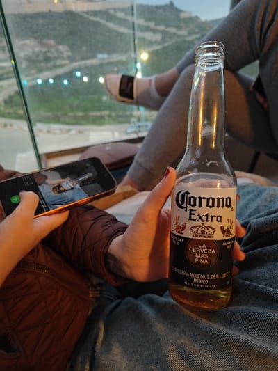 The image captures a relaxed indoor scene overlooking an outdoor landscape, likely during twilight or early evening. In the foreground, a person is holding a partially filled glass bottle of Corona Extra beer in their right hand, while simultaneously holding a smartphone horizontally in their left hand. The phone's screen displays what appears to be an image or video of a similar Corona Extra bottle, creating a self-referential visual. This person is wearing a brown quilted jacket and blue denim jeans; their hand shows red nail polish and a subtle tattoo on the wrist.

In the background, the lower legs and feet of a second person are visible, clad in light grey sweatpants and white socks with black accents, with one foot propped up. Both individuals appear to be in a comfortable, leisurely setting. A tall glass barrier or window separates the indoor space from the outdoors, revealing a hilly, somewhat arid landscape beyond. Distant bright lights, possibly from a road or buildings, dot the landscape, suggesting an elevated viewpoint overlooking a developed area. The sky above the hills has the soft, muted tones of dusk or dawn.

The text clearly visible on the beer bottle label reads: "Corona Extra", "LA CERVEZA MAS FINA" (The Finest Beer), "CERVECERIA MODELO, S. DE R.L. DE C.V.", "MEXICO", and "REG. S.S.A. N° 2417 "B"". The alcohol content "Alc. 4.6% beer" is also partially visible on the label.