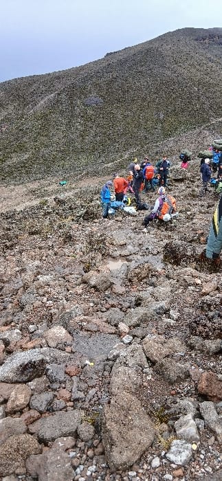 The scene at Gilman's Point, Tanzania, displays an outdoor, rocky landscape under an overcast sky. A steep, barren mountain slope covered in sparse, low-lying vegetation dominates the background. The foreground and mid-ground consist of dark, irregular volcanic rock formations and gravel, with multiple shallow puddles of brown water visible on the uneven ground. Approximately 15-20 individuals, dressed in cold-weather hiking apparel including insulated jackets in shades of blue, orange, red, black, and white, are clustered in the mid-ground. Many are equipped with large backpacks. Several individuals are standing, some are bent over appearing to adjust gear, and at least one person in an orange and white outfit is seated on the ground. Various pieces of equipment, including duffel bags and smaller items, are scattered near the groups. A partially visible individual in a green and yellow jacket is positioned in the lower right foreground. Small white and yellow objects are present on the rocky surface near the puddles.